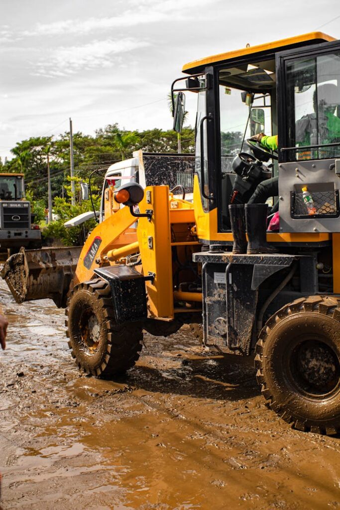 Back view of anonymous workman in modern loader with bucket on wet roadway with mud on sunny day